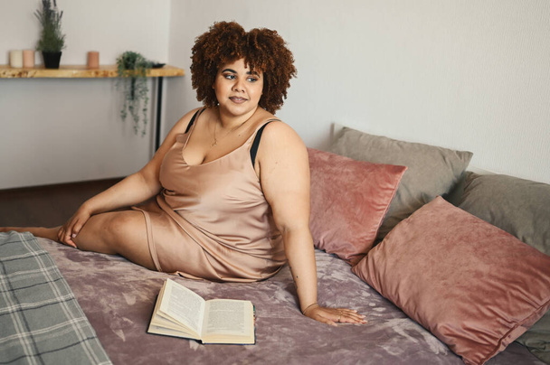 Beautiful curvy plus size African black woman afro hair lying on bed in silk powder pink dress cozy bedroom interior design. Body imperfection, body acceptance, body positive and diversity concept - Photo, Image