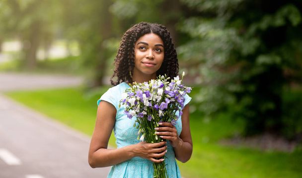 Fille noire rêveuse en robe romantique avec bouquet de fleurs au parc - Photo, image