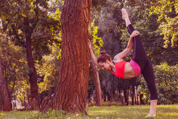 European girl in a light tracksuit doing yoga in the park in the afternoon in the summer. Gymnastics in nature. Thin woman outdoor. - 写真・画像