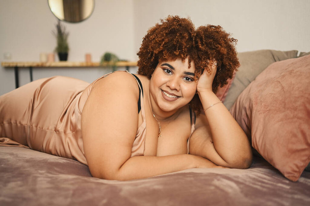 Piękny krzywy plus size Afrykański czarny kobieta afro włosy leżące na łóżku w jedwabiu pudrowy różowy sukienka przytulne wnętrza sypialni. Niedoskonałość ciała, akceptacja ciała, koncepcja ciała pozytywnego i różnorodności - Zdjęcie, obraz