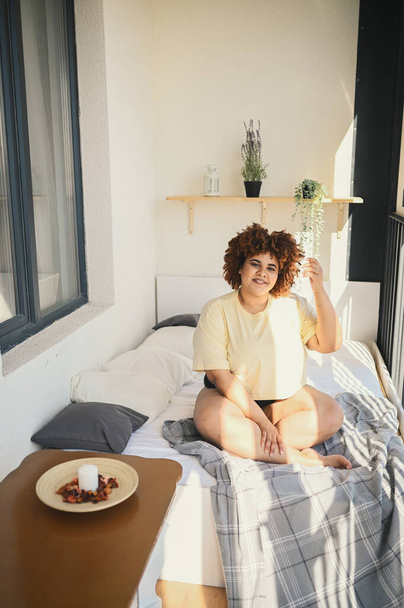 Beautiful happy curvy plus size African black woman afro hair posing in beige t-shirt and underwear on sunny balcony bedroom. Body imperfection, body acceptance, body positive and diversity concept - Photo, Image
