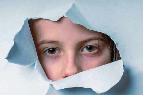 Portrait of young cute scared child girl looking through a hole in the paper wall. Close up. - Photo, Image