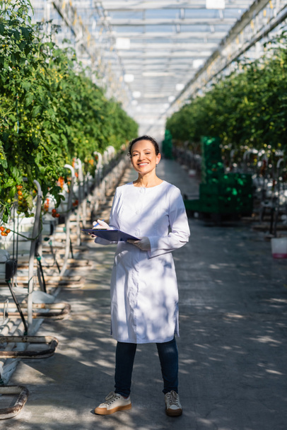 happy african american quality inspector standing with clipboard in greenhouse - Photo, Image