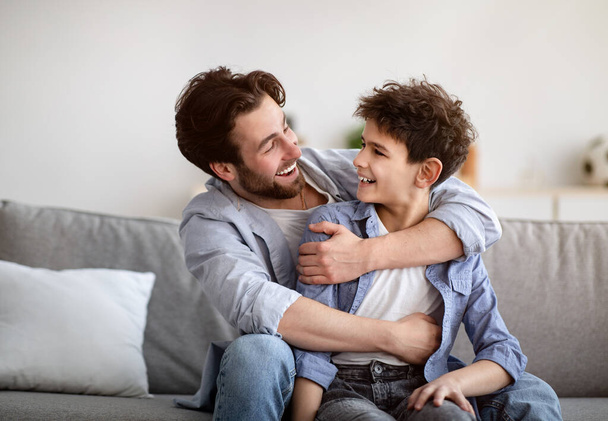 Concepto de dos generaciones. Feliz padre abrazando al hijo, mirándose y sonriendo, sentado en el sofá en casa - Foto, Imagen
