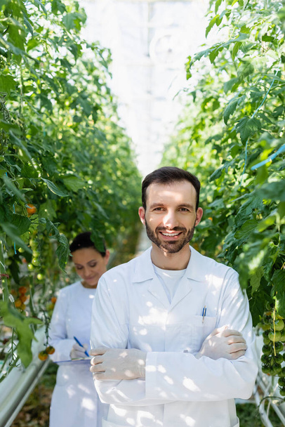 usmívající se zemědělský technik při pohledu na kameru poblíž afrického amerického kolegy pracující na rozmazaném pozadí - Fotografie, Obrázek