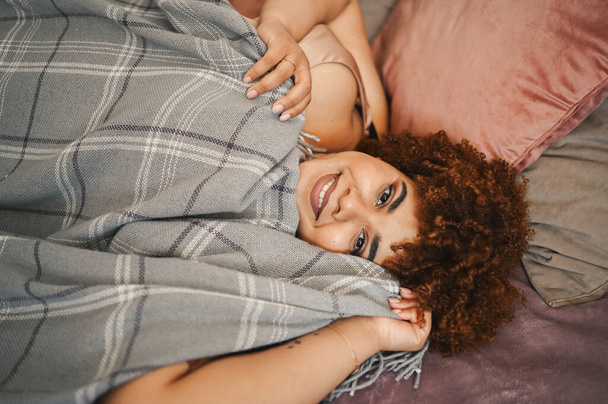Beautiful curvy plus size African black woman afro hair lying on bed with grey plaid blanket cozy bedroom interior design. Body imperfection, body acceptance, body positive and diversity concept - Photo, Image