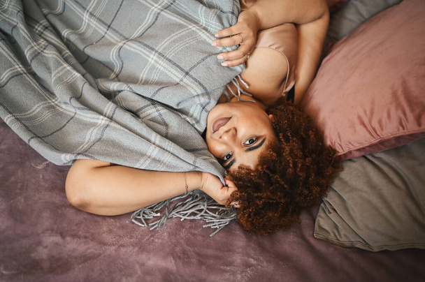 Beautiful curvy plus size African black woman afro hair lying on bed with grey plaid blanket cozy bedroom interior design. Body imperfection, body acceptance, body positive and diversity concept - Foto, immagini