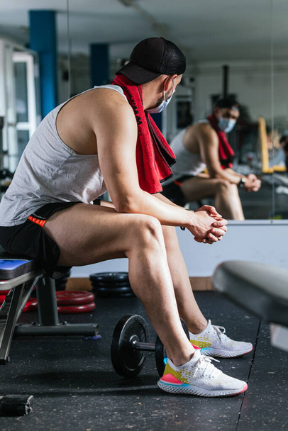 Ο άνθρωπος κάθεται στο γυμναστήριο με μια πετσέτα γύρω από το λαιμό του. Περιτριγυρισμένο από μηχανές και βάρη. Έννοια υγείας και ευεξίας - Φωτογραφία, εικόνα