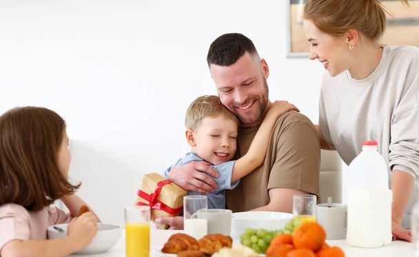 Ο καλύτερος μπαμπάς. Χαριτωμένο χαρούμενο αγοράκι που αγκαλιάζεται με τον μπαμπά, δίνοντάς του τυλιγμένο κουτί δώρου για την ημέρα των πατέρων ή των γενεθλίων, ενώ τρώει πρωινό με την οικογένεια στην κουζίνα στο σπίτι. Οικογενειακές διακοπές - Φωτογραφία, εικόνα