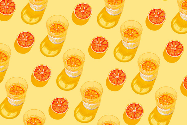 Kreatives Muster aus geschnittener Grapefruit, Blutorange und Glas mit Limonade oder Wasser auf gelbem Hintergrund. Sommerfrucht- und Erfrischungskonzept. Minimaler Stil. Sonnenlicht flach lag. Ansicht von oben - Foto, Bild