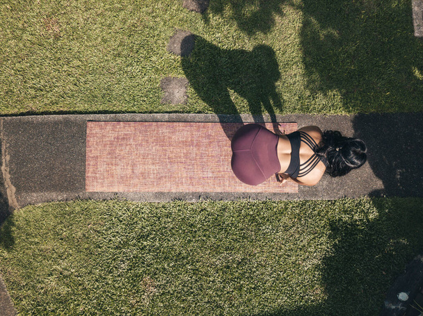 Вид сверху на женщину, делающую упражнения на растяжку при подготовке к сеансу йоги. На тропинке в парке или саду дома. - Фото, изображение