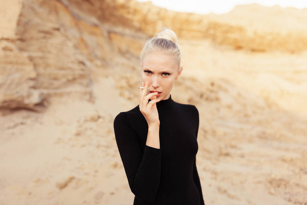 Confidence young woman with blond hair and makeup, holds a cigarette and smokes, looking at camera, against sandy career, at sunset. Front portrait. - Фото, изображение