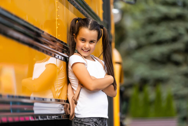 Χαριτωμένο κορίτσι με ένα σακίδιο στέκεται κοντά στο λεωφορείο πηγαίνει στο σχολείο ποζάροντας στην κάμερα σκεπτική γκρο πλαν - Φωτογραφία, εικόνα