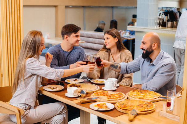 A group of young cheerful friends is sitting in a cafe talking and eating pizza. Lunch at the pizzeria. - Фото, изображение