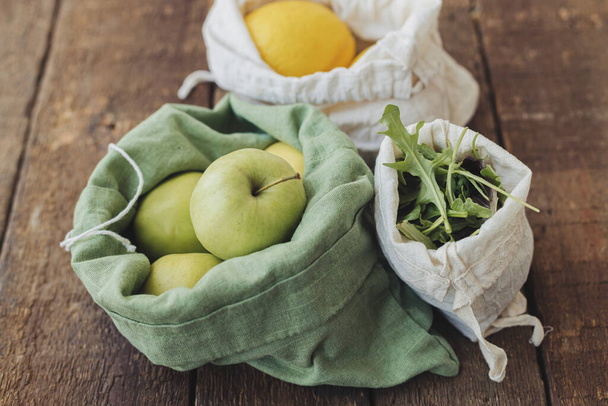 Plastic free grocery shopping. Fresh apples, lemons and arugula in eco cotton bags on rustic wooden table. Zero waste. Organic fruits and vegetables in reusable bags. Sustainable lifestyle - Foto, imagen