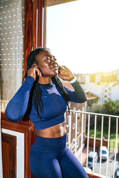schwarze Afro-Frau in Sportkleidung, die mit Kopfhörern in einem Fenster ihres Hauses Musik hört, weil sie aufgrund der Covid19 Coronavirus-Pandemie zu Hause Sport treiben wird - Foto, Bild