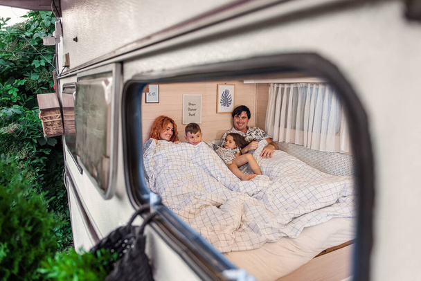 Cheerful family of travelers lie on the bed in mobile home, trailer house. People are having fun, mother father smiling at children. Brother sister fooling around, happily spending weekend at picnic - Photo, Image