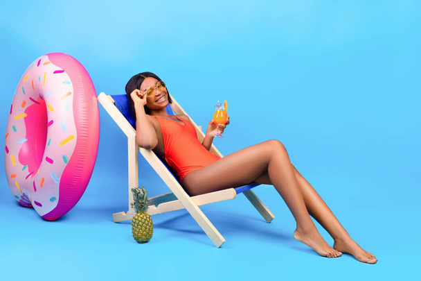 Portrait complet de jeune femme noire se refroidissant dans une chaise longue avec un délicieux cocktail tropical sur fond bleu - Photo, image