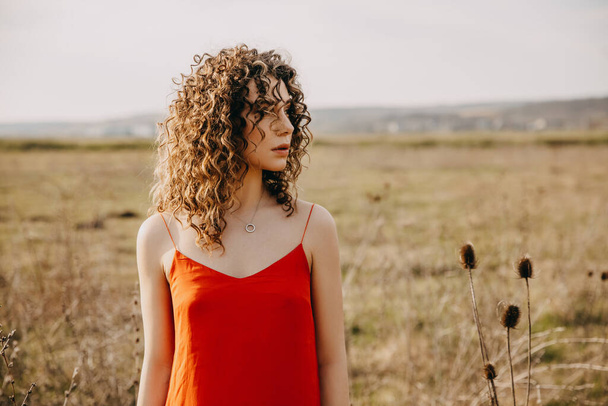 Joven mujer tranquila con el pelo rizado, con un vestido rojo, al aire libre en un campo. - Foto, imagen