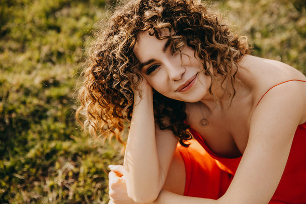 glückliche junge brünette Frau mit lockigem Haar, lächelnd, im Freien, auf einem Feld, in die Kamera blickend. - Foto, Bild