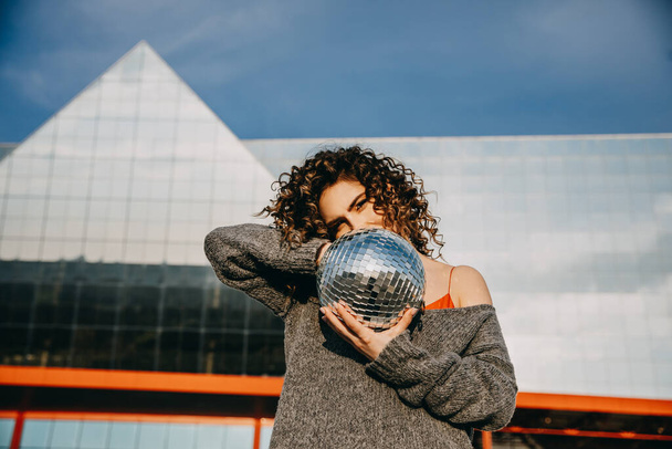Portrait of a young woman with curly hair, holding a disco ball, outdoors, on city background. - Foto, Bild