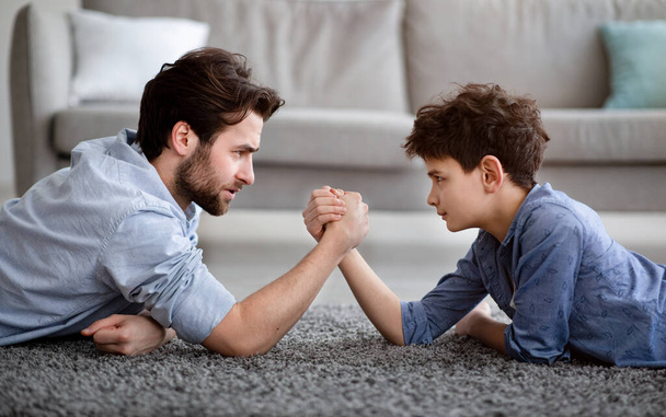 Raising a real man. Concentrated father and son arm wrestling and competing while lying on floor - Photo, Image