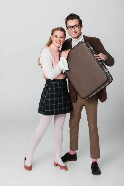 счастливая пара в ретро-одежде улыбается в камеру, держа винтажный чемодан на сером фоне - Фото, изображение