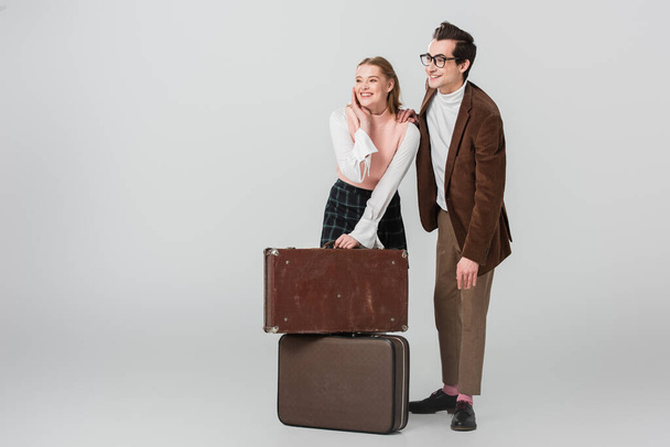 joyful couple in retro outfit looking away near suitcases on grey background - Photo, Image