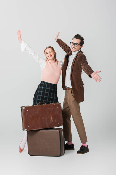 cheerful couple standing with open arms near vintage suitcases on grey background - Fotó, kép