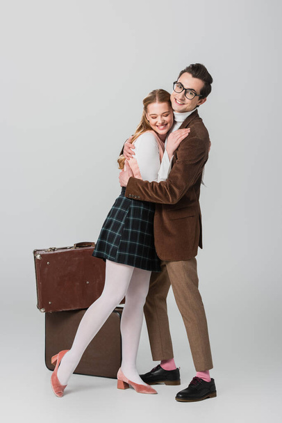 happy couple in retro outfit embracing near suitcases on grey background - Photo, Image