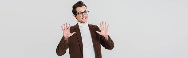 homme joyeux dans les lunettes et blazer brun posant avec les paumes ouvertes isolé sur gris, bannière - Photo, image