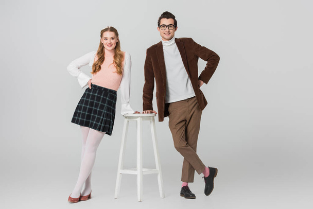 стильная пара в винтажном наряде, позирующая с руками на бедрах возле высокого стула на сером фоне - Фото, изображение
