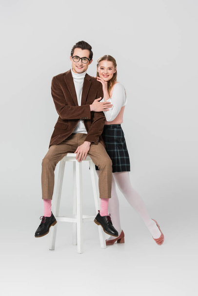 happy woman in plaid skirt leaning on shoulder of man sitting on high stool on grey background - Foto, Imagem