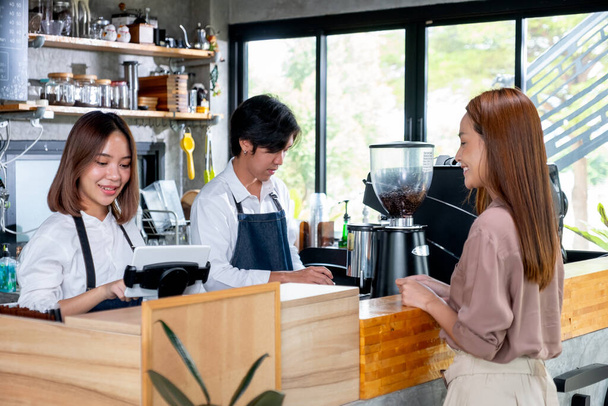 Aasialainen barista tai kahvinkeitin saa tilauksen asiakasnaiselta, kun taas hänen työkaverinsa valmistaa kahvia koneella. Hyvän tiimityön käsite tukee pienyritysjärjestelmää. - Valokuva, kuva