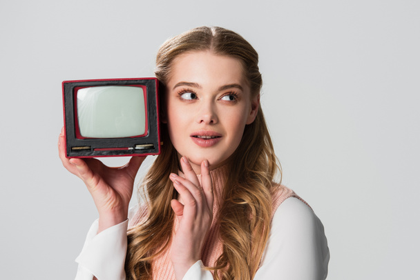 dreamy woman touching chin and looking away while holding small vintage tv isolated on grey - Fotoğraf, Görsel