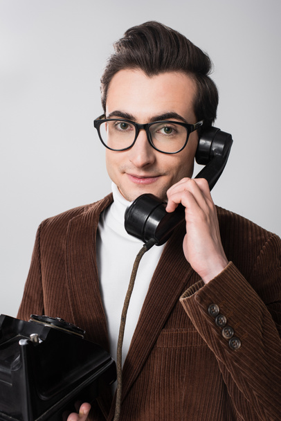 jovem homem em óculos olhando para a câmera enquanto fala no telefone vintage isolado em cinza - Foto, Imagem
