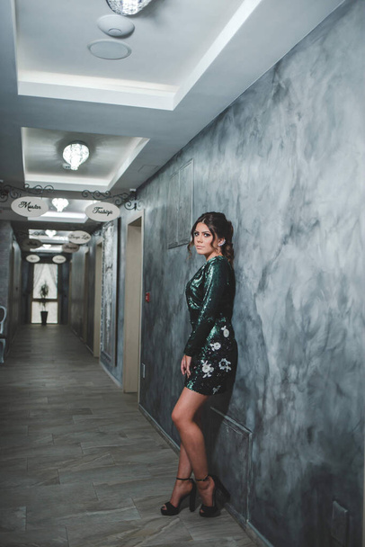 An adult woman wearing a green sparkling dress with high heels and leaning on a restaurant hallway wall - Fotoğraf, Görsel