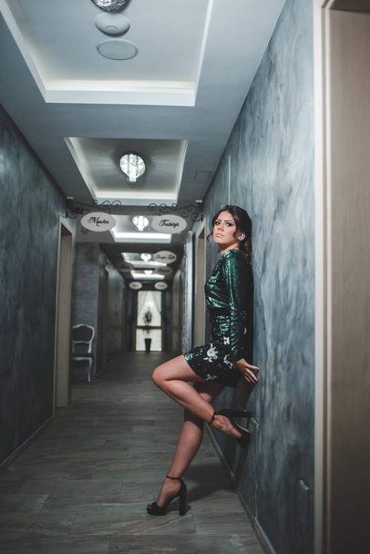 An adult woman wearing a green sparkling dress with high heels and leaning on a restaurant hallway wall - Photo, image