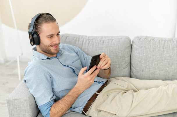 Satisfied handsome guy using smartphone and wireless headphones for listening music - Фото, изображение