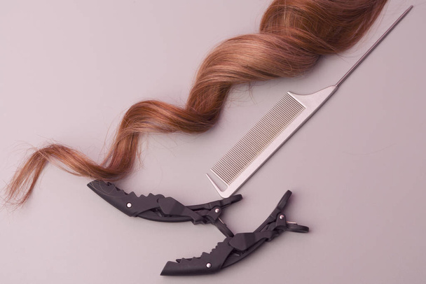 Curly Strand of Ginger Hair with Metallic Grey Tail Comb and Black Hair Clip on a Warm Light Gray Background, Copy Space. Professional Salon Tools, Example of Artificial Hair - Photo, Image