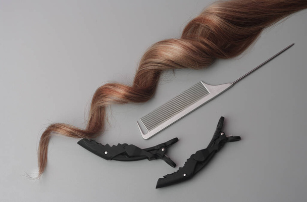 Curly Lock of Blond Hair with Metallic Grey Tail Comb and Black Hair Clip on a Gray Background, Αντιγραφή χώρου. Επαγγελματικά εργαλεία σαλόνι, Παράδειγμα τεχνητής τρίχας - Φωτογραφία, εικόνα