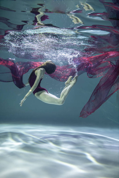 όμορφο κορίτσι με κόκκινο μαγιό με κόκκινο παρεό κάτω από το νερό σε μπλε φόντο  - Φωτογραφία, εικόνα