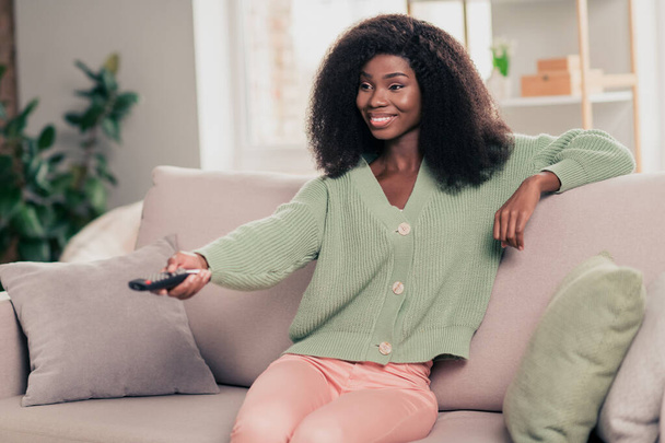 Photo portrait of smiling curly girl watching tv sitting on couch at home keeping remote controller - Foto, Imagen