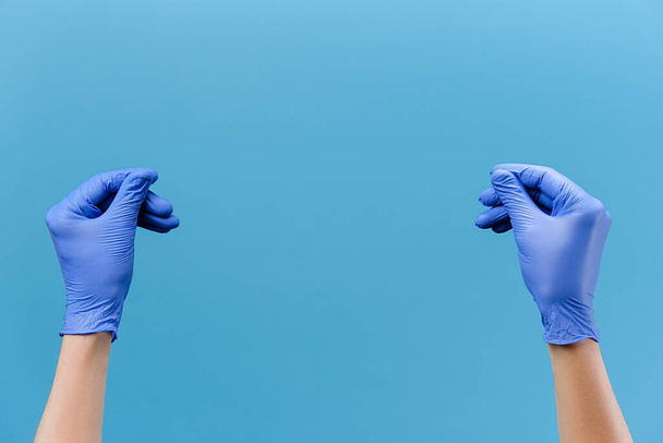 Close up of male hands in medical protective latex gloves rubbing fingers showing cash gesture asking for money, isolated on blue studio wall with copy space for advertisement. Body language concept - Foto, Imagem