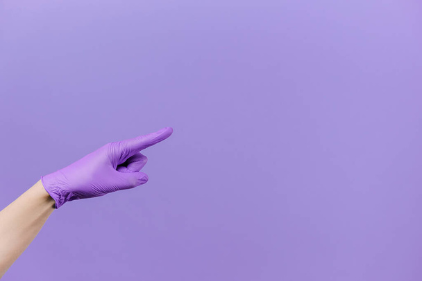 Male finger pointing at copy space, wears medical protective latex gloves, isolated on purple studio background wall with copy space for advertisement. Advertising area, mockup. Hand gesture concept - Photo, Image