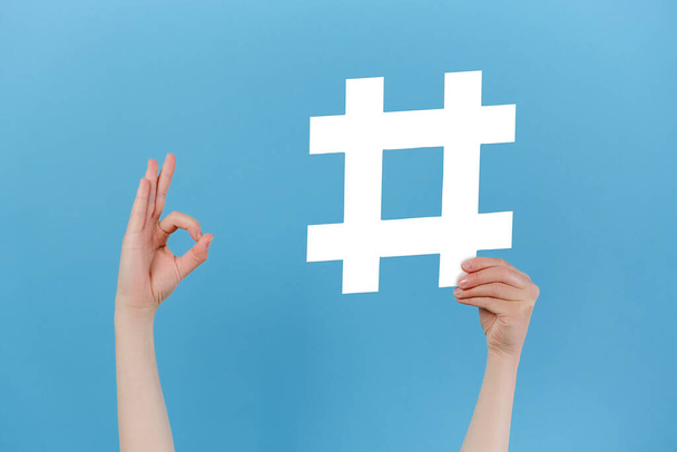 Unrecognizable young female holding large big hashtag sign and showing okay gesture, viral web content, internet promotion, isolated on pastel blue studio wall. Blogging blog website strategy concept - Photo, image