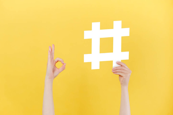 Close up of unrecognizable female holding large big hashtag sign and showing okay gesture, viral web content, internet promotion, isolated on pastel yellow wall. Blogging blog website strategy concept - Фото, изображение