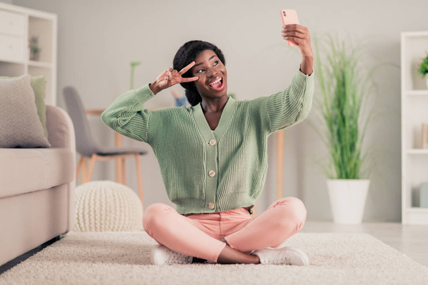 Full body photo of young excited afro girl happy positive smile make selfie smartphone show peace cool v-sign sit floor home - Foto, afbeelding