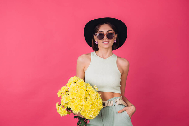 Beautiful stylish woman in hat and sunglasses posing over pink red background holding large bouquet of yellow asters, spring mood, positive emotions isolated space - Photo, Image