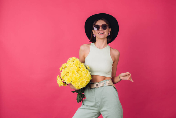 Beautiful stylish woman in hat and sunglasses posing over pink red background holding large bouquet of yellow asters, spring mood, positive emotions isolated space - Photo, Image